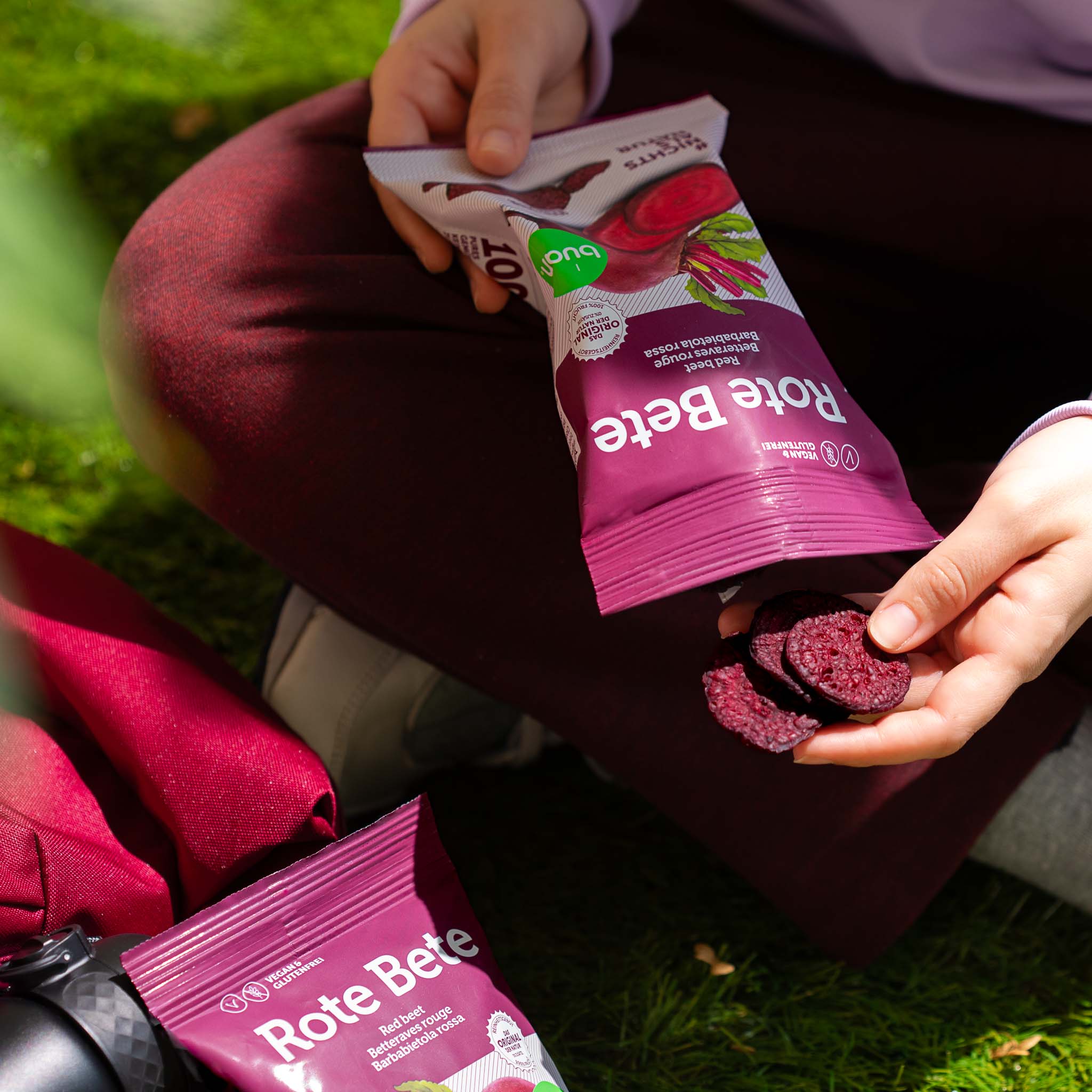 Person im Schneidersitz nimmt einige vakuumgetrocknete Rote Bete Chips aus einer Packung buah Gemuesechips