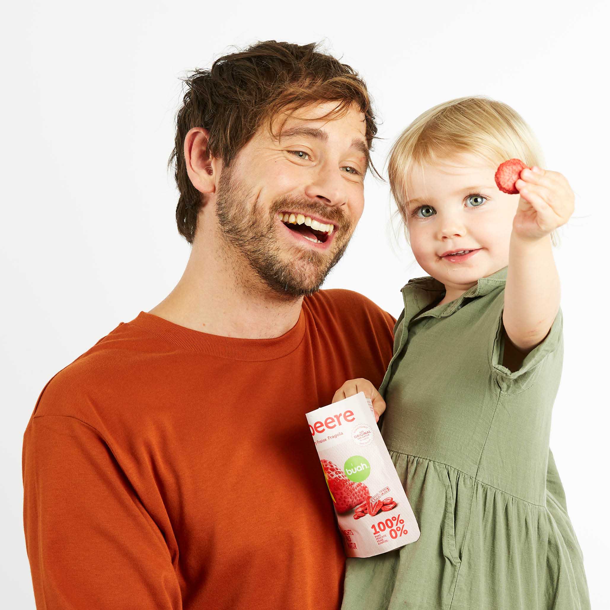 Kleines Kind bei Papa auf dem Arm hält ein Snackpack buah Erdbeere in der Hand und lacht