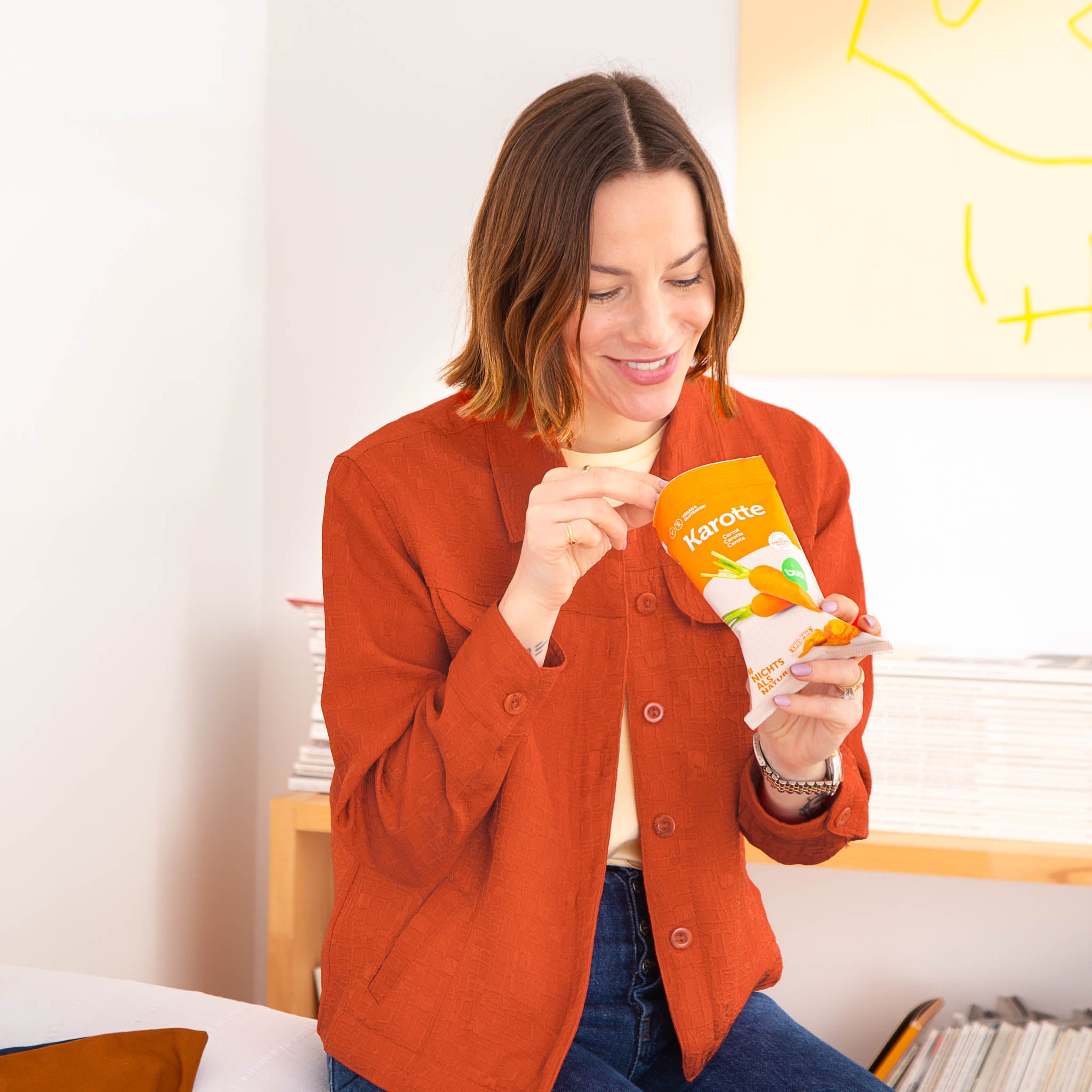 Lachende Frau mit rostrotem Blazer isst vakuumgetrocknete Karottenchips aus einer Packung buah Karottenchips