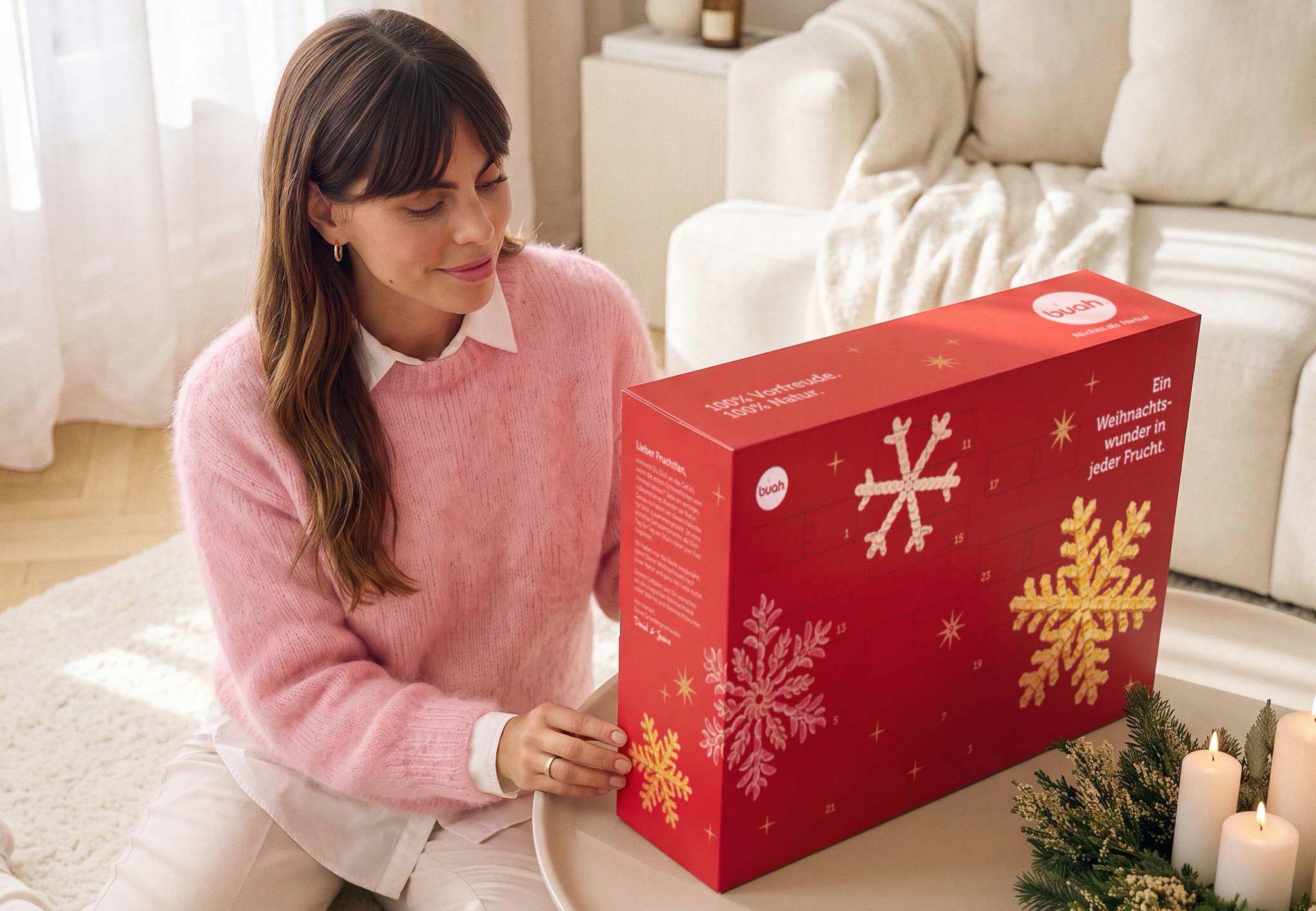 Eine lächelnde Frau im rosa Pullover sitzt auf dem Boden an einem Couchtisch und betrachtet voller Vorfreude den roten buah Adventskalender. Sie ist dabei, eines der Türchen zu öffnen.