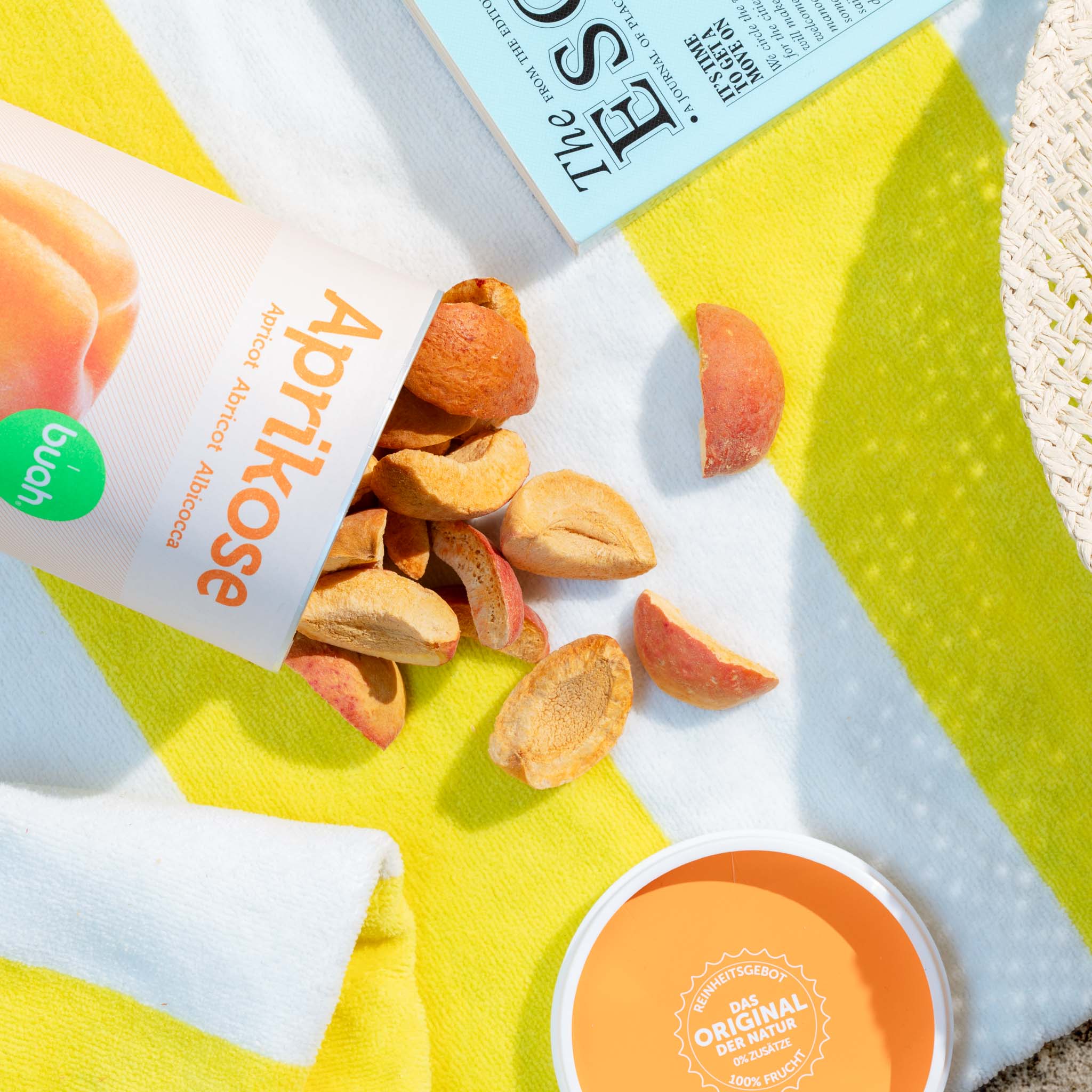 Boîte d'abricots lyophilisés buah renversés sur une serviette de plage jaune et blanche, l'abricot repose sur la serviette à côté du livre