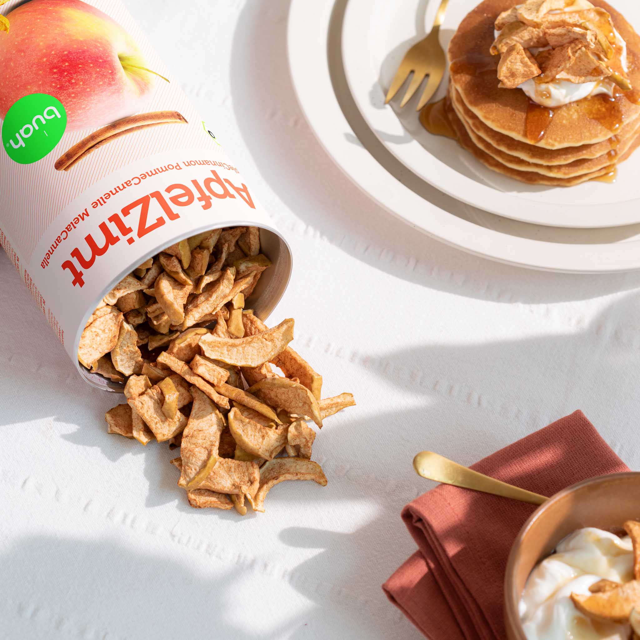 Tisch mit weisser Tischdecke, Teller mit Apfel-Pancakes, Bowl mit Joghurt, daneben eine umgekippte Dose buah ApfelZimt, die knackigen Stuecke fallen aus der Dose und liegen auf dem Tisch