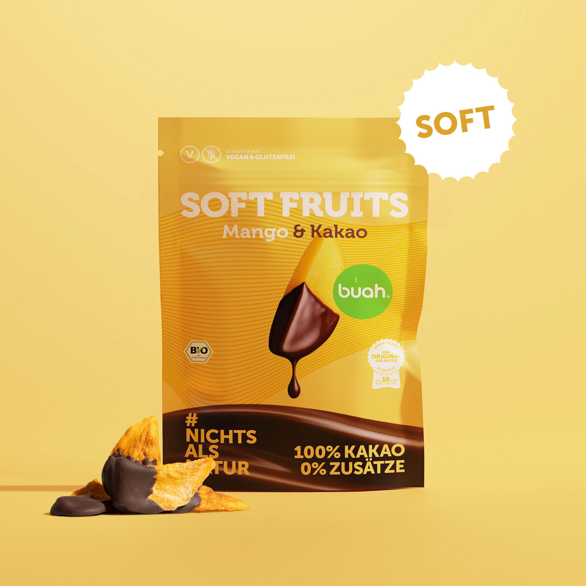 Ein gelber Beutel buah Soft Fruits der Sorte Mango & Kakao steht vor einem gelben Hintergrund. Davor liegen einige softe, mit Schokolade überzogene Mango-Stücke.