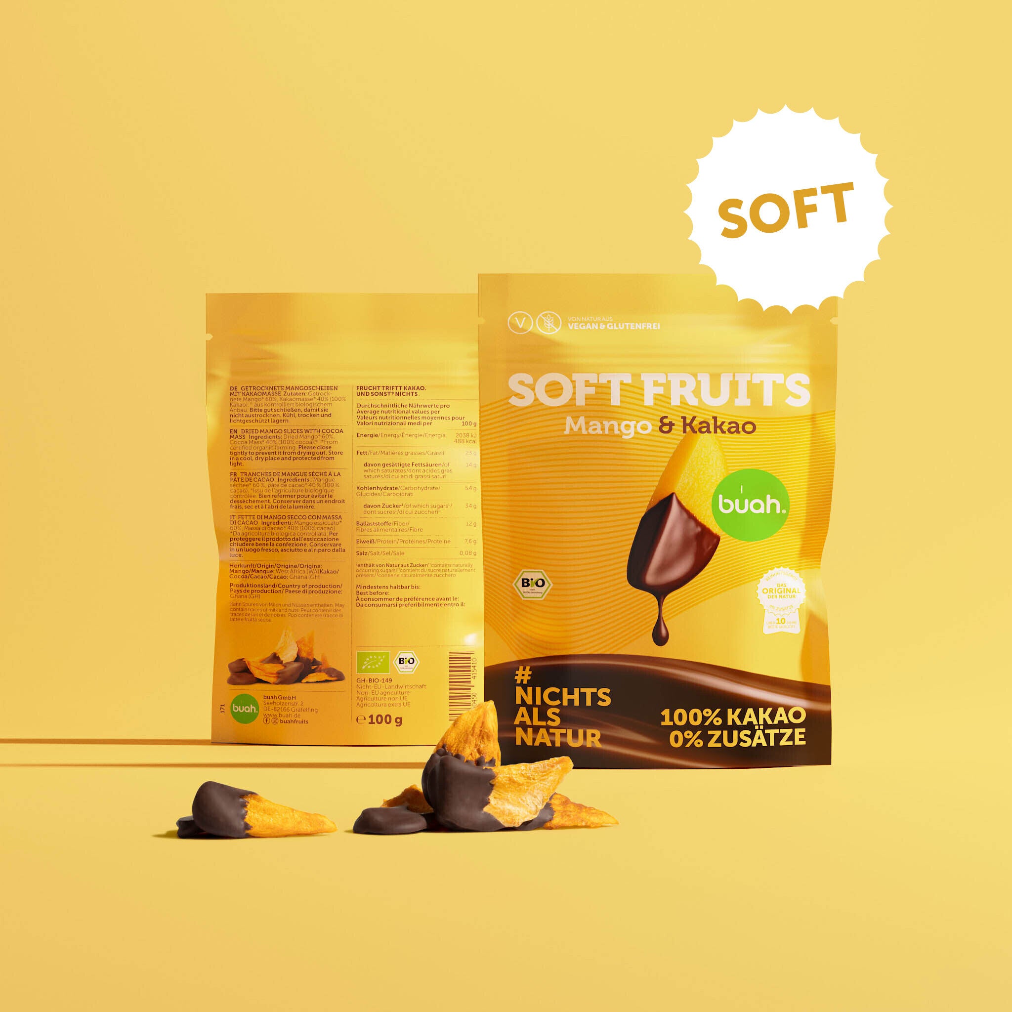 Ein gelber Beutel buah Soft Fruits der Sorte Mango & Kakao steht vor einem gelben Hintergrund. Dahinter ist die Rückseite der Verpackung zu erkennen. Davor liegen einige softe, mit Schokolade überzogene Mango-Stücke.