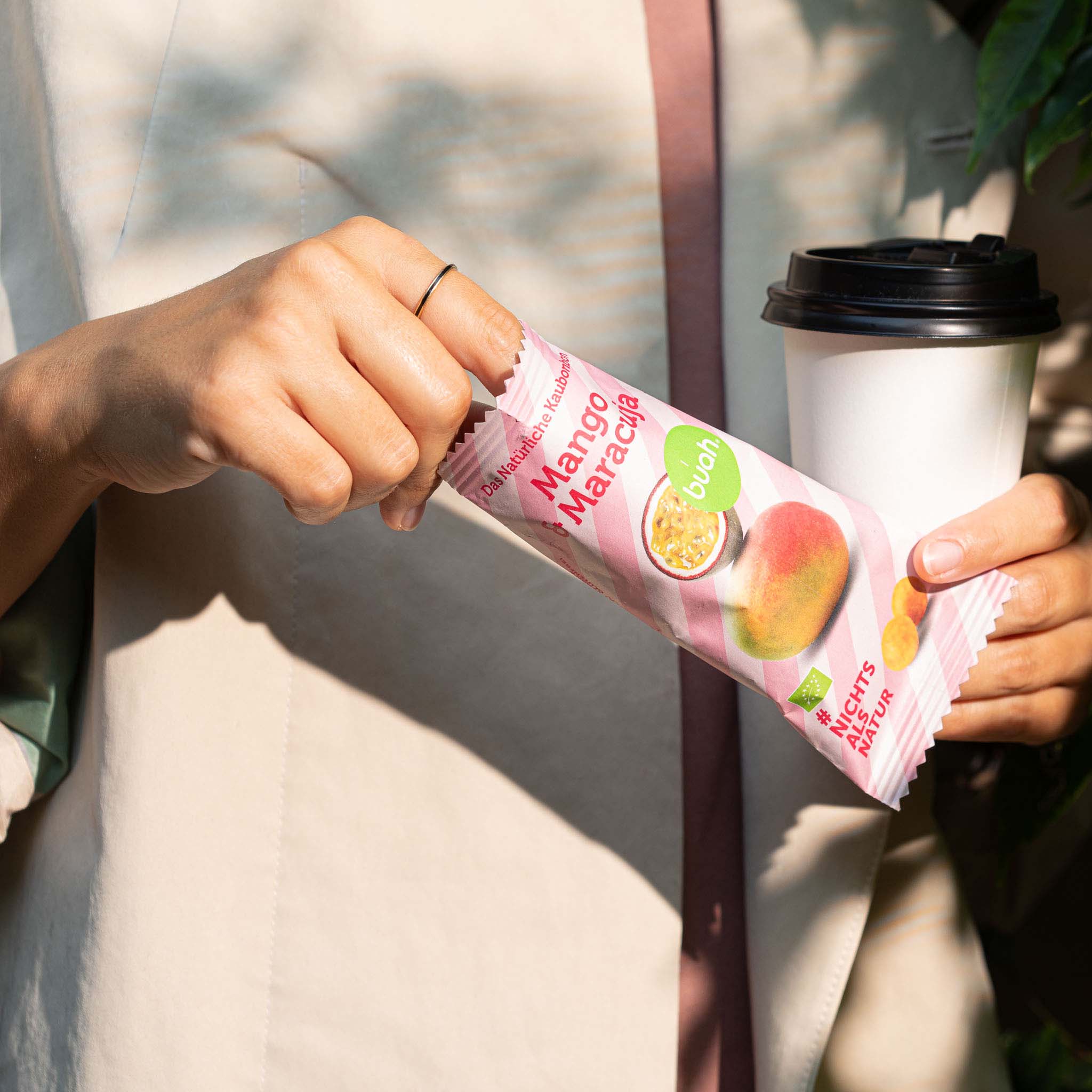 Person hält Kaffeebecher und nimmt ein Mango-Maracuja Kaubonbon von buah aus der Verpackung