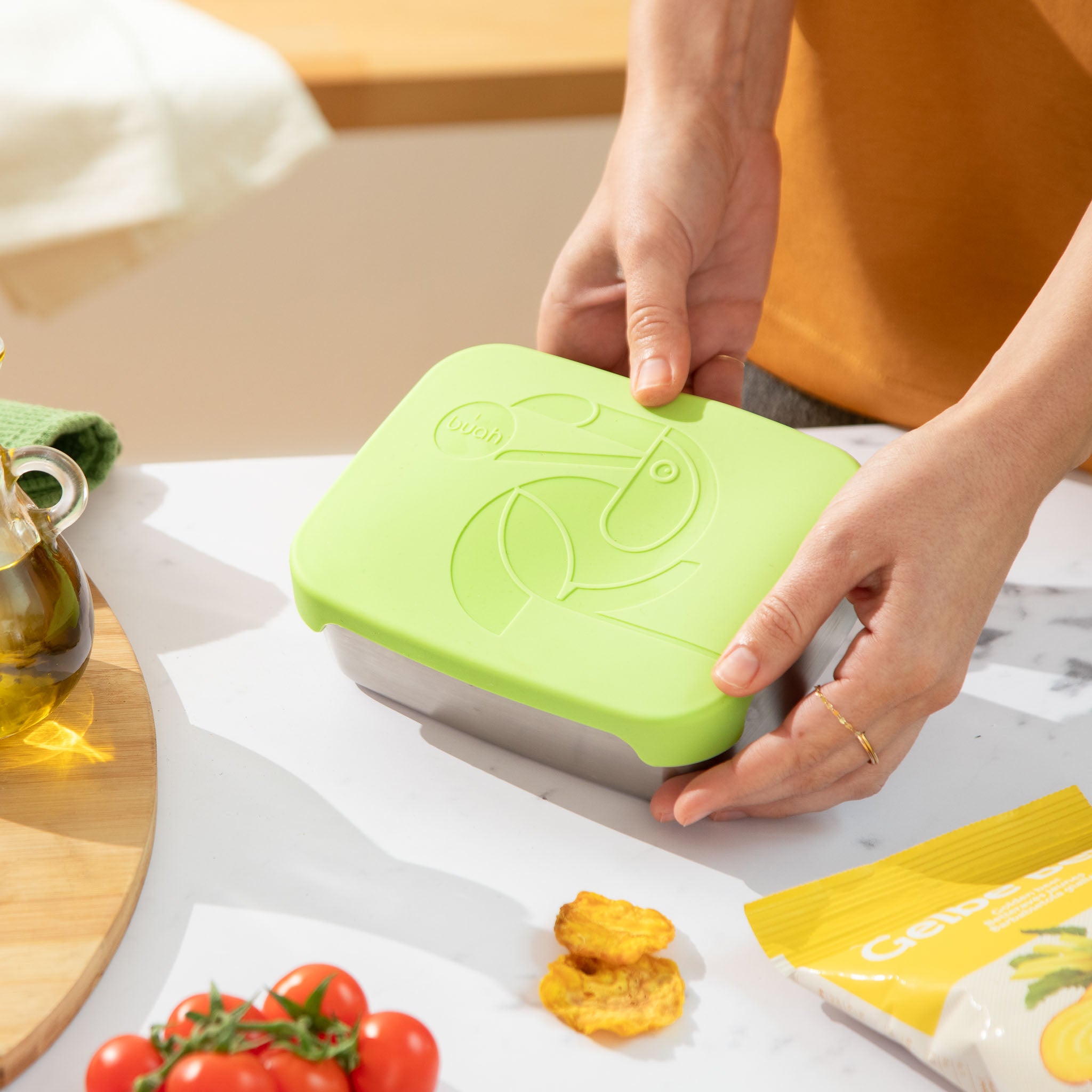 buah Brotdose aus Edelstahl mit gruenem Deckel aus Silikon liegt auf einem Esstisch und wird von einer Person hochgehalten.