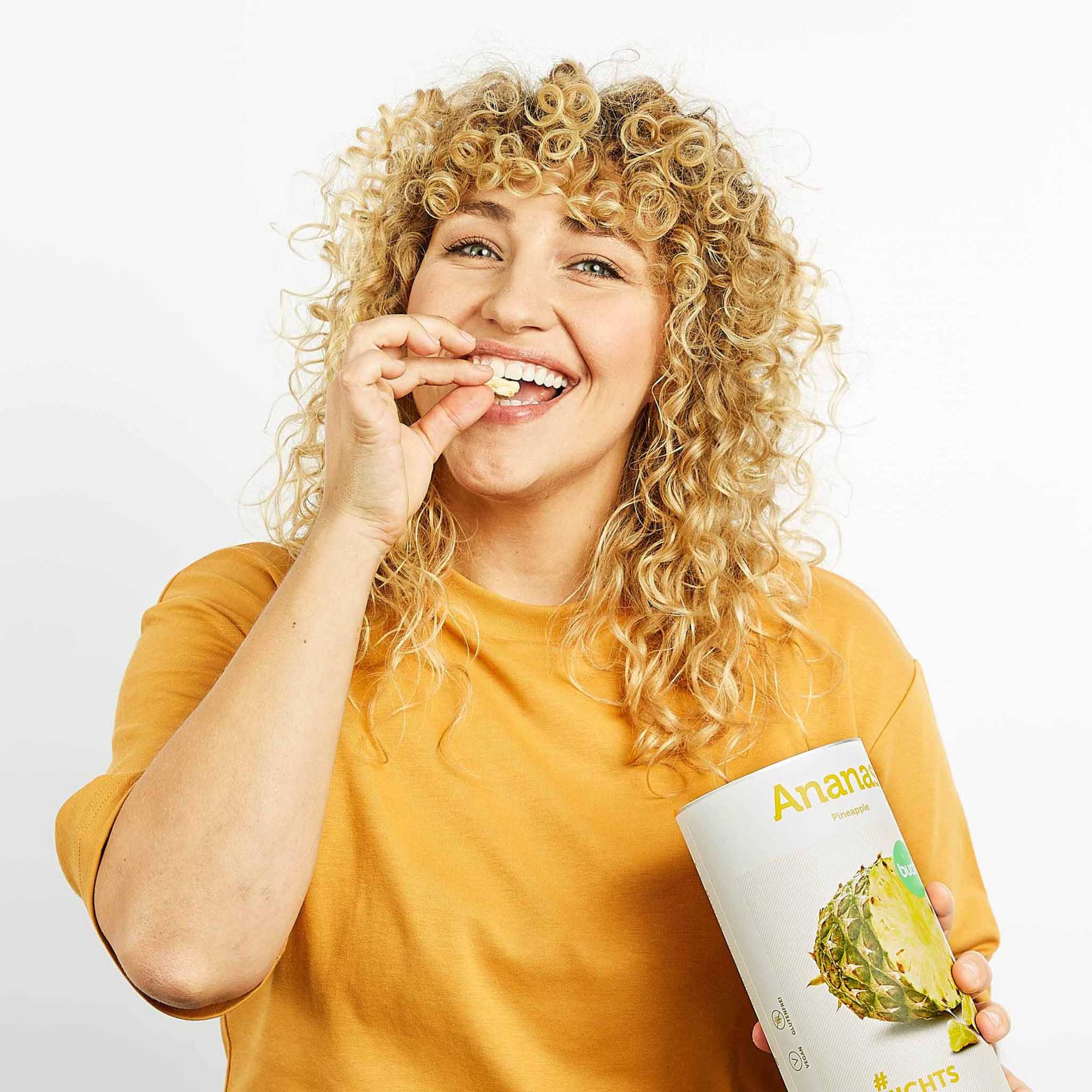 Lachende Frau mit blonden Locken hat eine Dose buah Ananas in der Hand und isst die gefriergetrocknete Ananas