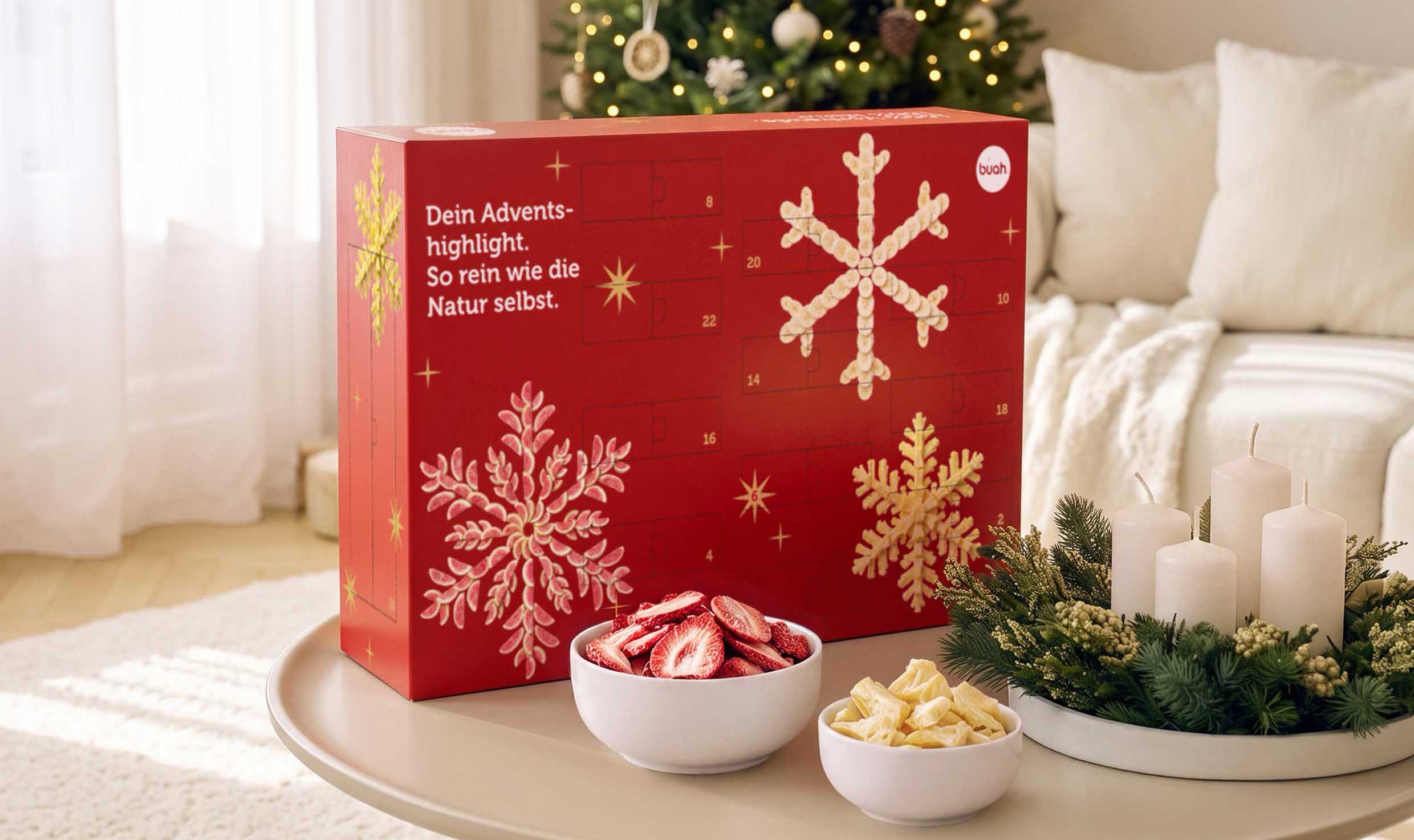 Ein roter buah Adventskalender steht auf einem Wohnzimmertisch vor einem geschmückten Weihnachtsbaum. Auf dem Kalender sind Schneeflocken aus Fruchtstücken abgebildet. Davor stehen zwei weiße Schälchen mit gefriergetrockneten Erdbeeren und hellen Fruchtstücken sowie festliche weiße Kerzen.