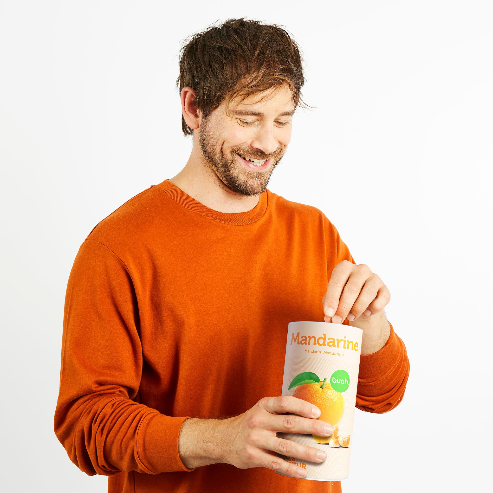 Mann mit braunen Haaren und orangenem Pullover hält eine Dose buah Mandarinen in der Hand