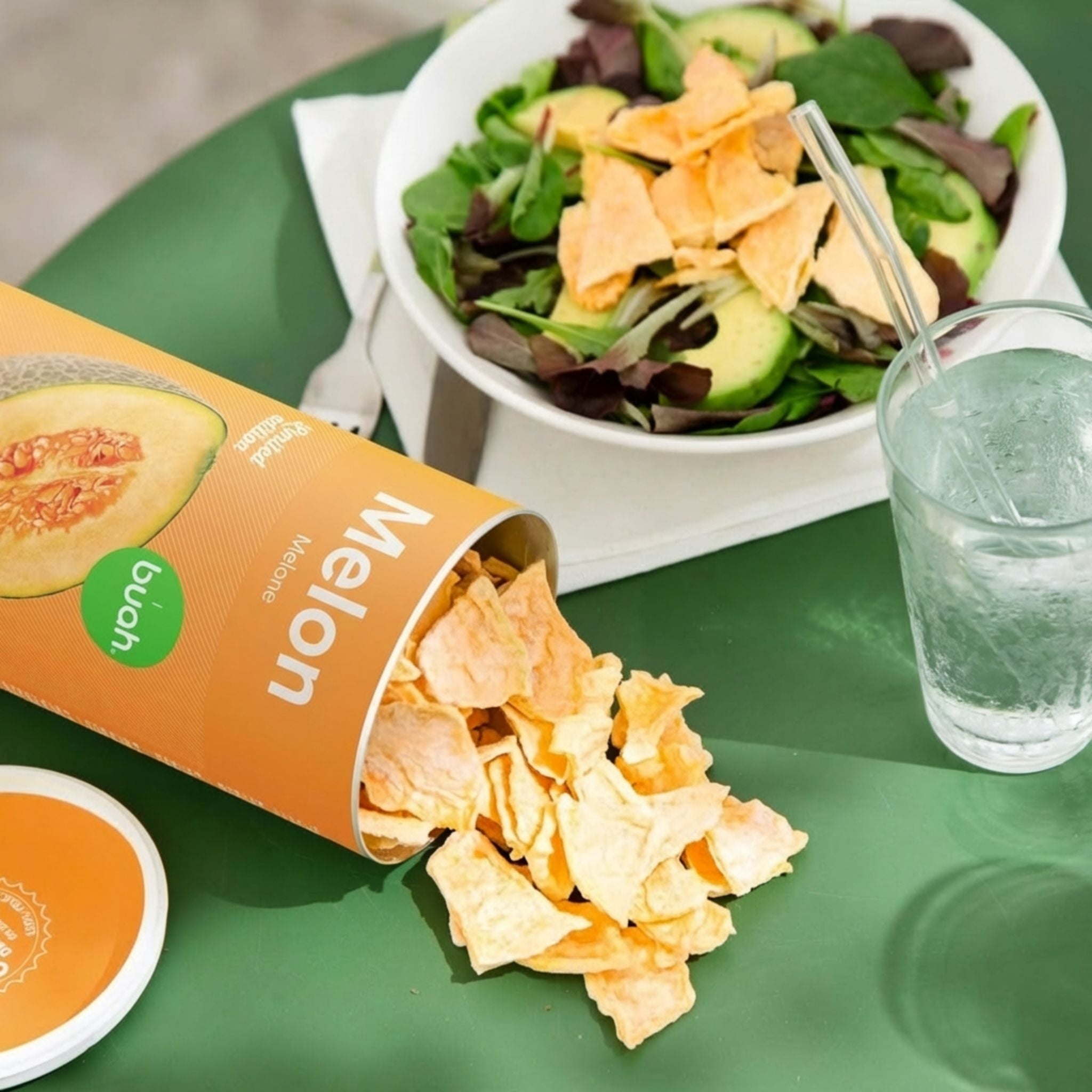 Une boîte de fruits lyophilisés buah à la saveur melon est couchée sur une table de jardin verte, avec des morceaux de fruits qui en sortent. En arrière-plan, on aperçoit une salade fraîche et un verre d'eau.