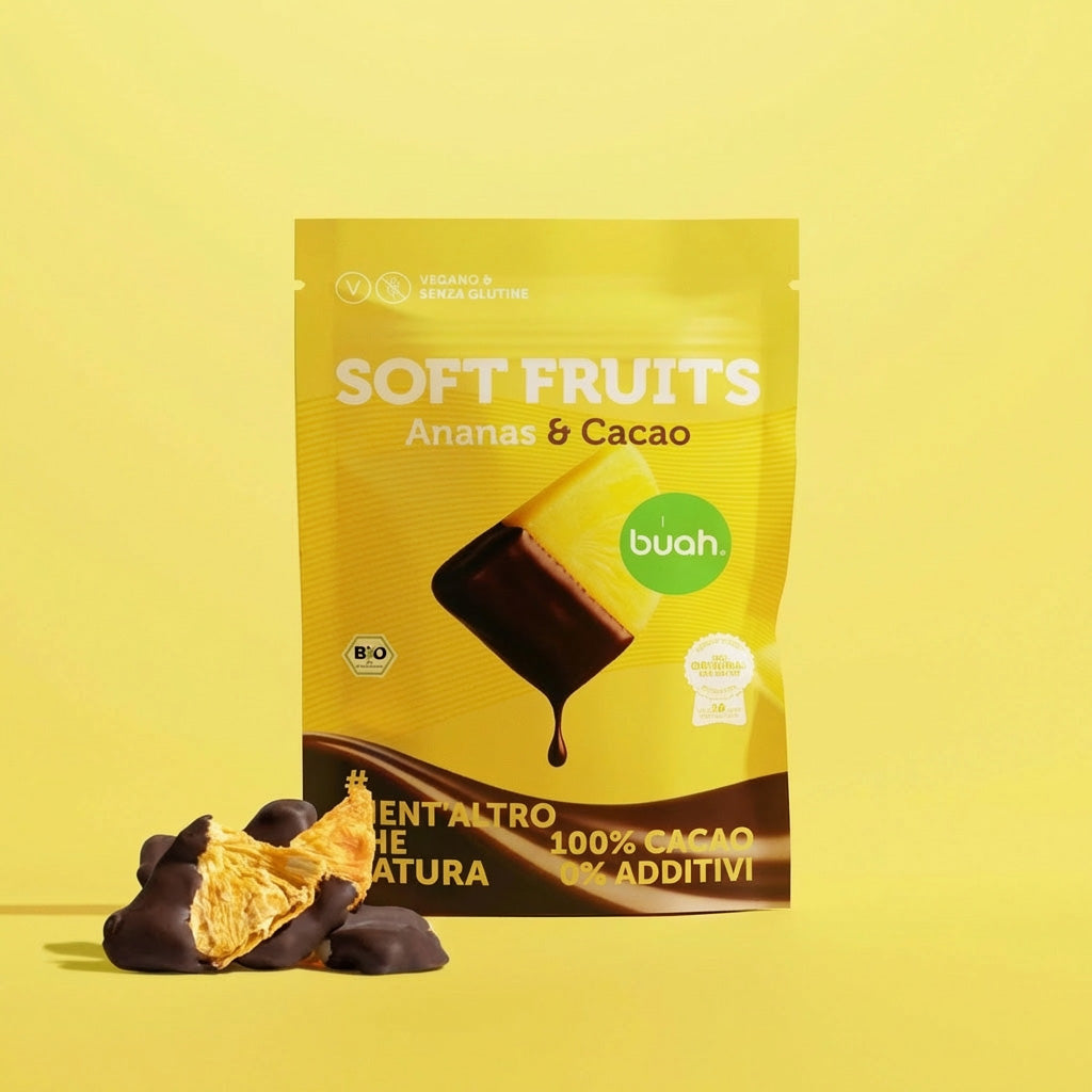 Un sacchetto giallo di buah Soft Fruits al gusto Ananas & Cacao si trova su uno sfondo giallo. Davanti ci sono alcuni morbidi pezzi di ananas ricoperti di cioccolato.