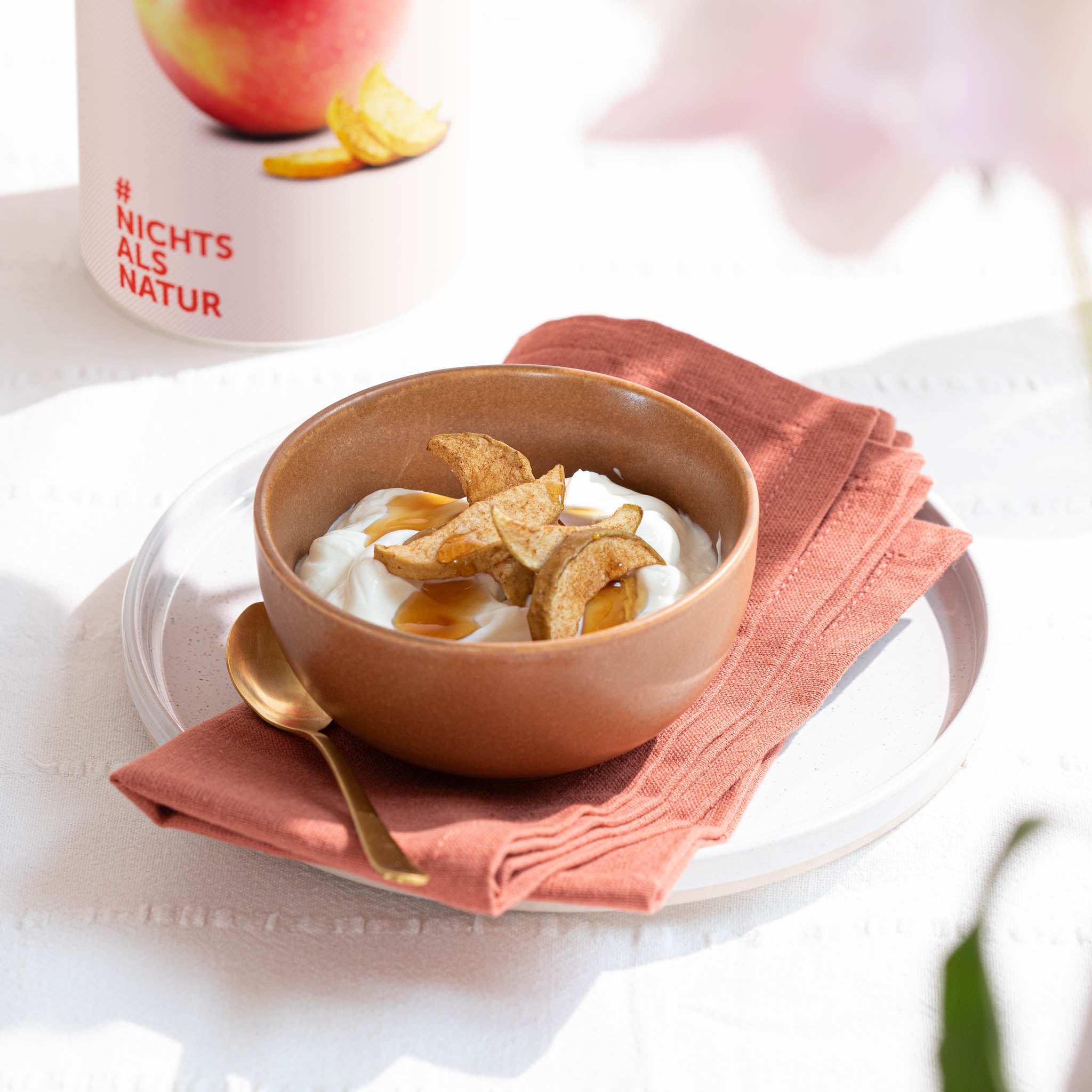 Braune Bowl steht auf einer lachsfarbenen Serviette auf einem Teller, Inhalt Joghurt mit Honig und winterlichem, knusprigen buah ApfelZimt, goldener Loeffel daneben