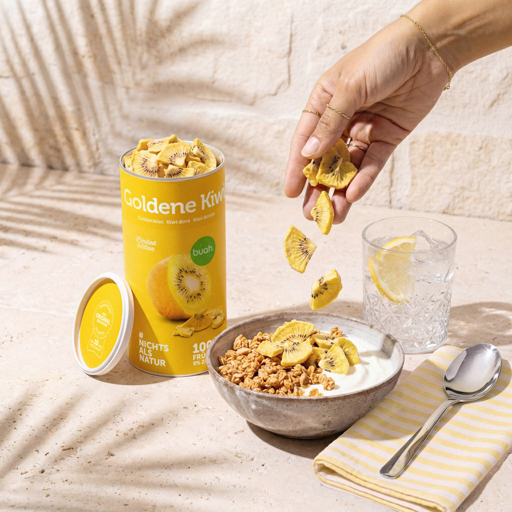 Eine sommerliche Szene auf einer hellen Steinplatte. Eine Hand streut knusprige buah gefriergetrocknete Kiwi-Stücke aus einer gelben Dose über eine Schale mit Müsli und Joghurt. Daneben steht ein Glas erfrischendes Wasser mit einer Zitronenscheibe.