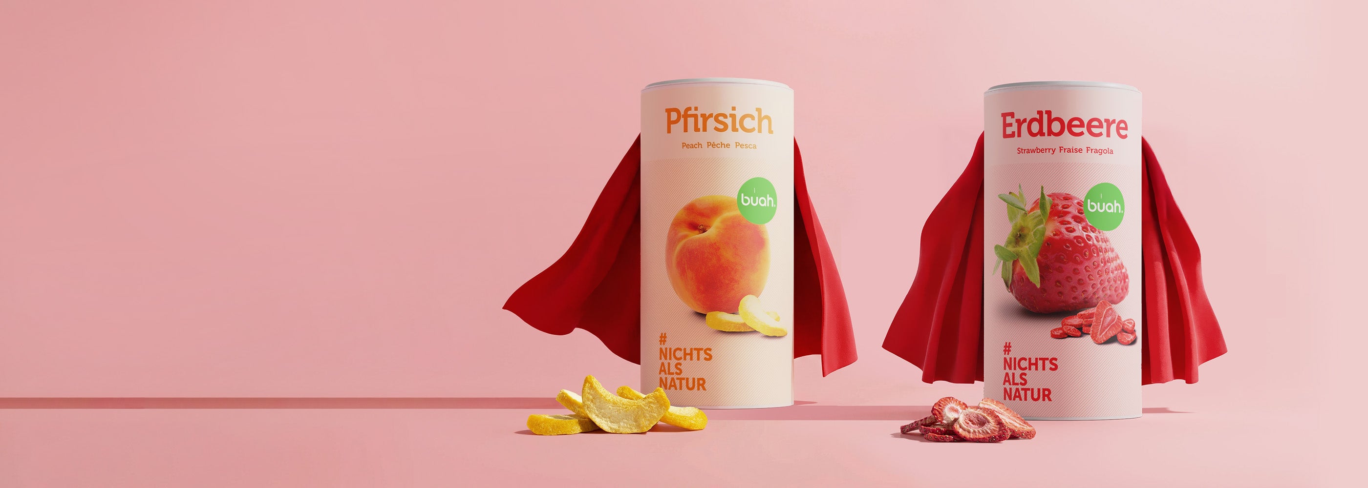 Ein breites Banner mit zwei großen Dosen buah gefriergetrocknete Früchte der Sorten Pfirsich und Erdbeere vor einem pinken Hintergrund. Beide Dosen tragen rote Superhelden-Umhänge. Vor den Dosen liegen Fruchtstücke. Die linke Bildhälfte besteht aus einer großen Freifläche.