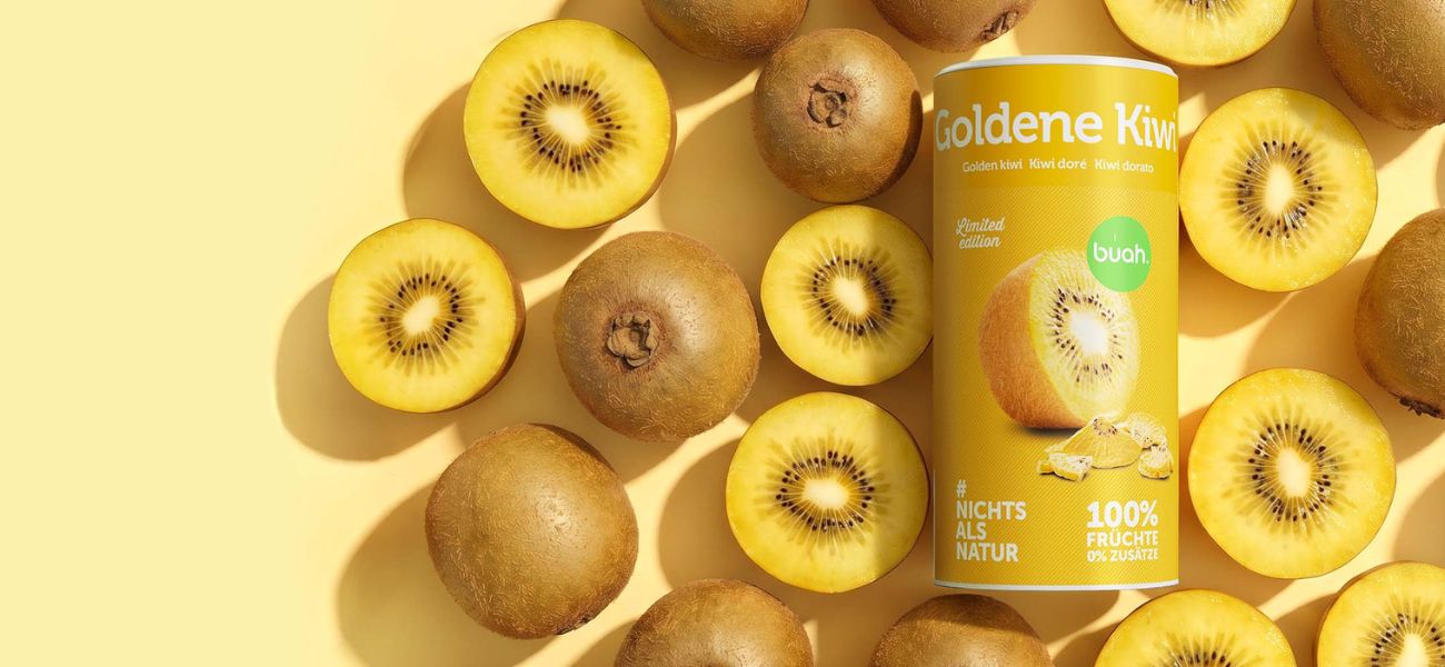Eine flach von oben fotografierte Anordnung im Querformat auf hellgelbem Hintergrund. Im Zentrum steht eine gelbe Dose buah gefriergetrocknete Goldene Kiwi (Limited Edition), umgeben von zahlreichen frischen, ganzen und halbierten Goldenen Kiwis.