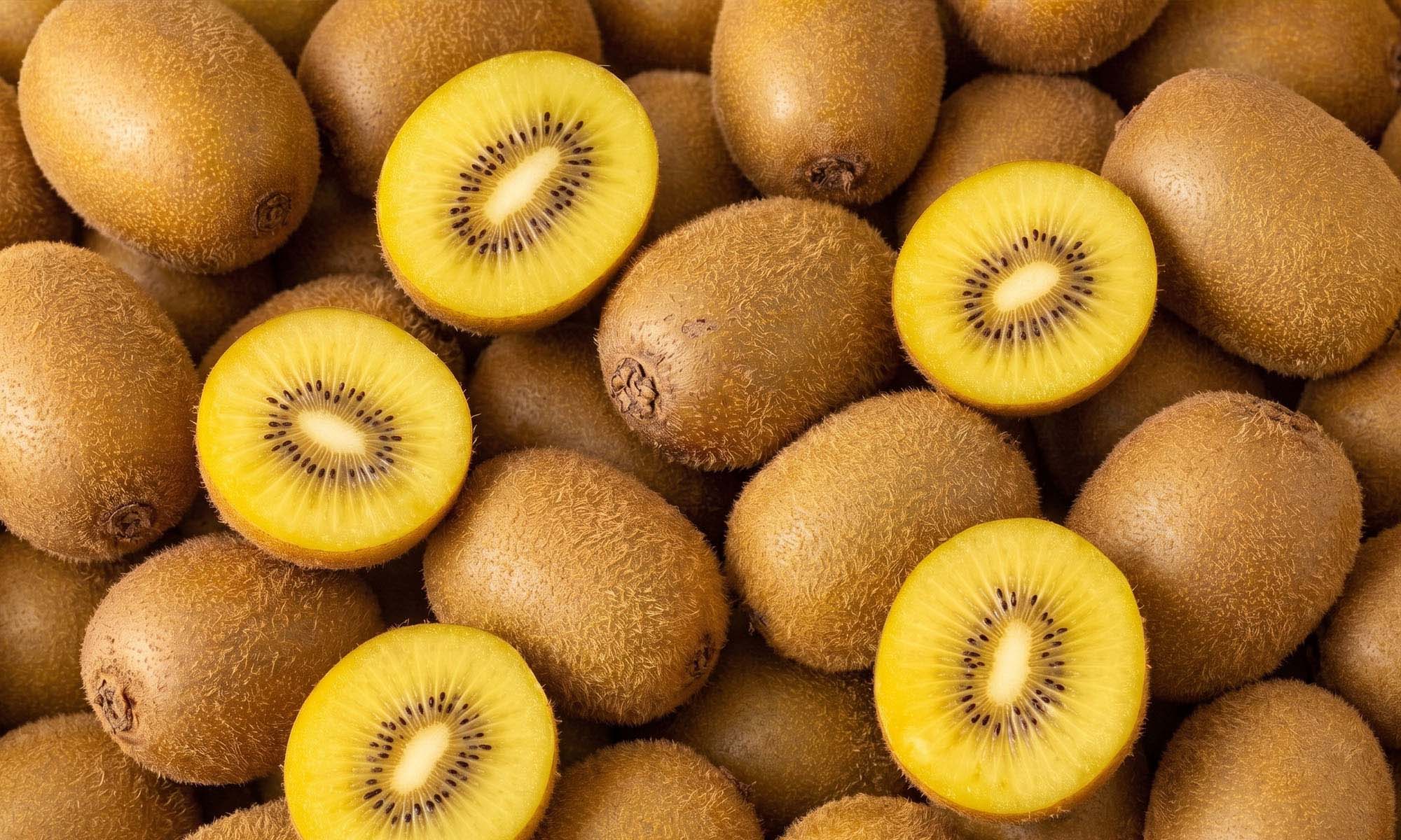 Eine strukturierte Nahaufnahme von vielen frischen Goldenen Kiwis, die dicht an dicht liegen. Einige halbiert Früchte zeigen das saftige, leuchtend gelbe Fruchtfleisch mit den schwarzen Kernen.