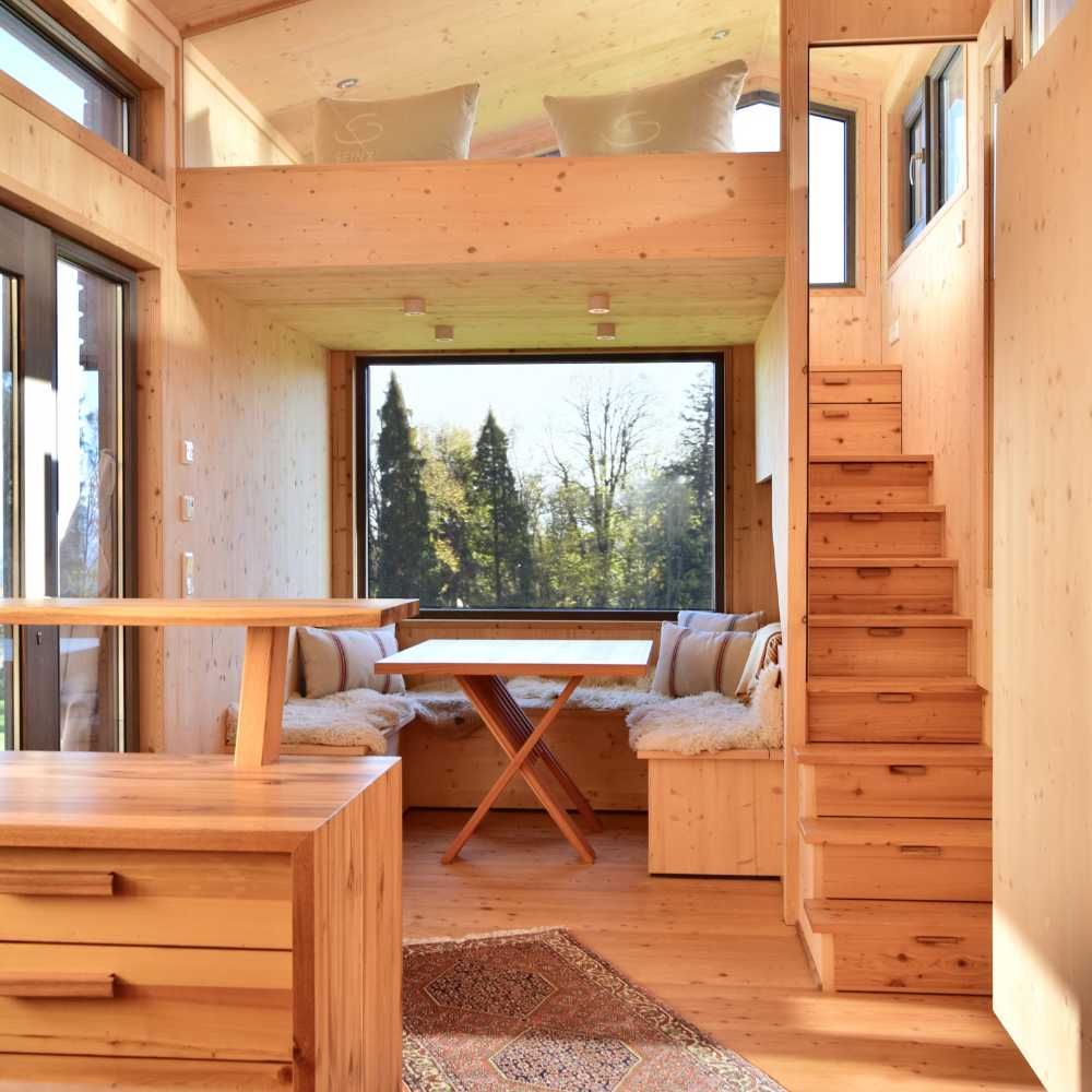 Das helle, gemütliche Innere einer modernen Holzhütte aus hellem Holz. Eine Sitzecke mit Holztisch und flauschigen Kissen steht vor einem großen Panoramafenster mit Blick in die Natur. Rechts führt eine Holztreppe hinauf.